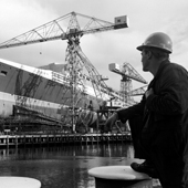 Photograph of three quarter bow view of the Cunard liner RMS Queen Elizabeth 2, with ship worker in foreground, taken from west side of dock during construction at the shipyard of John Brown and Company, Clydebank, 30 Aug 1967 (Crown Copyright, National Records of Scotland, UCS1/118/736/1155)